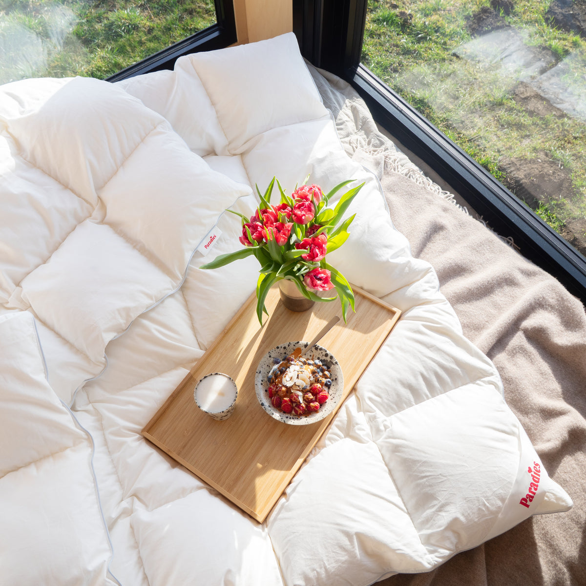 Draufsicht auf gemütliches Bett mit weißer Daunendecke, Holztablett mit Tasse, Teller mit Beeren und Vase mit roten Tulpen vor großem Fenster
