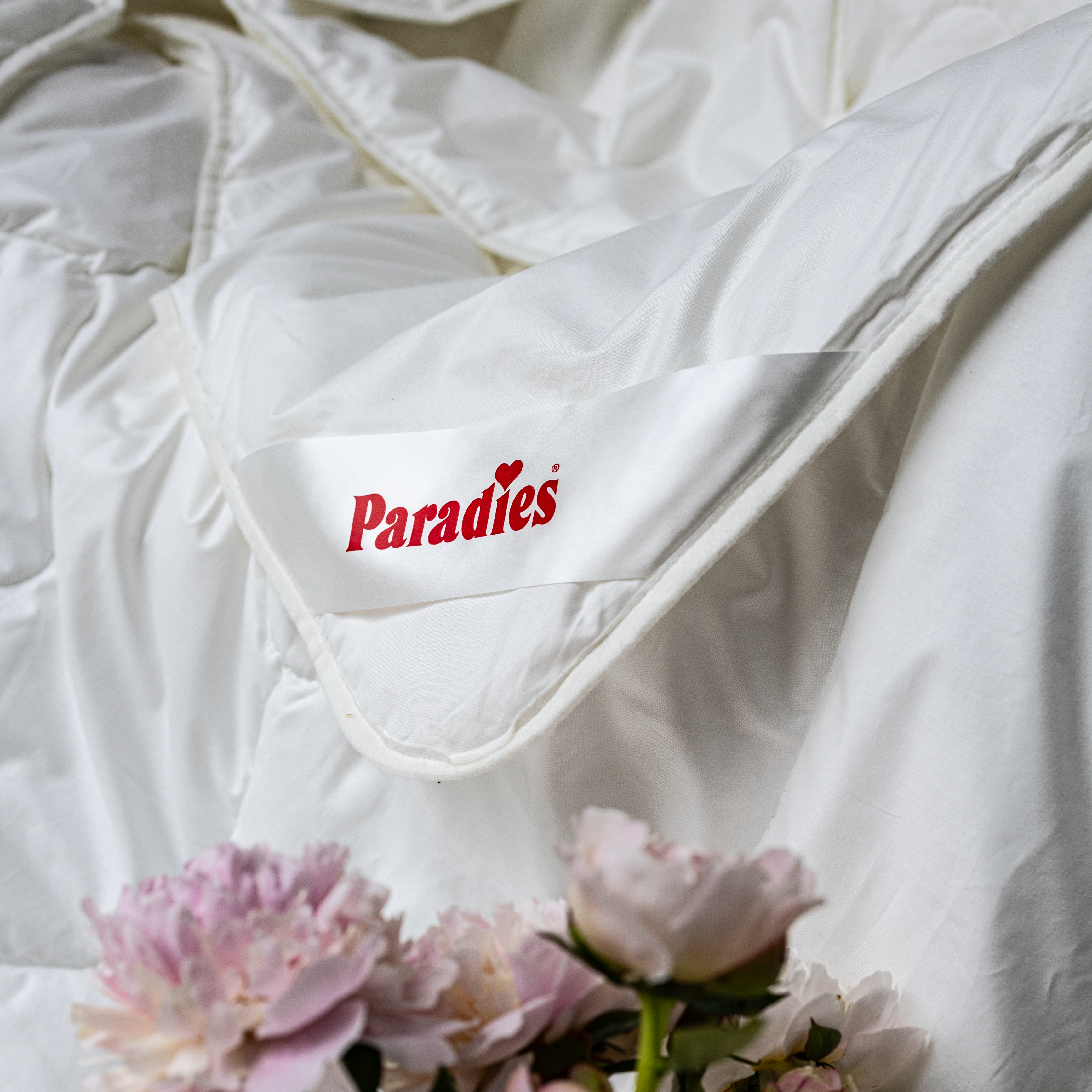 Detailaufnahme des Ecklabels der Paradies Prima Sommerbettdecke, umgeben von rosa Pfingstrosenblüten