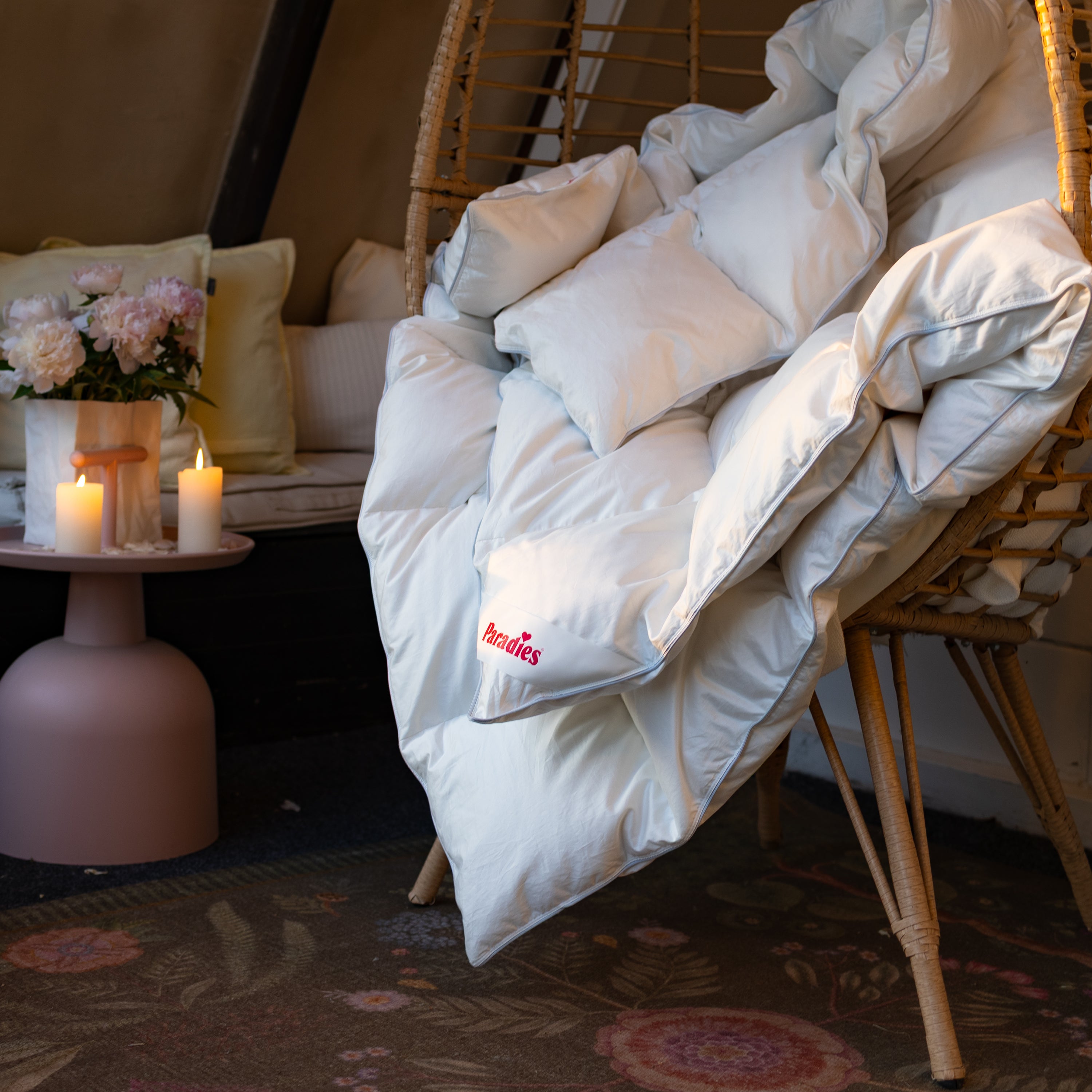 Weiße Paradies Ganzjahresbettdecke Safina 100 liegt locker gefaltet in einem geflochtenen Sessel neben einem Tisch mit brennenden Kerzen und Blumen