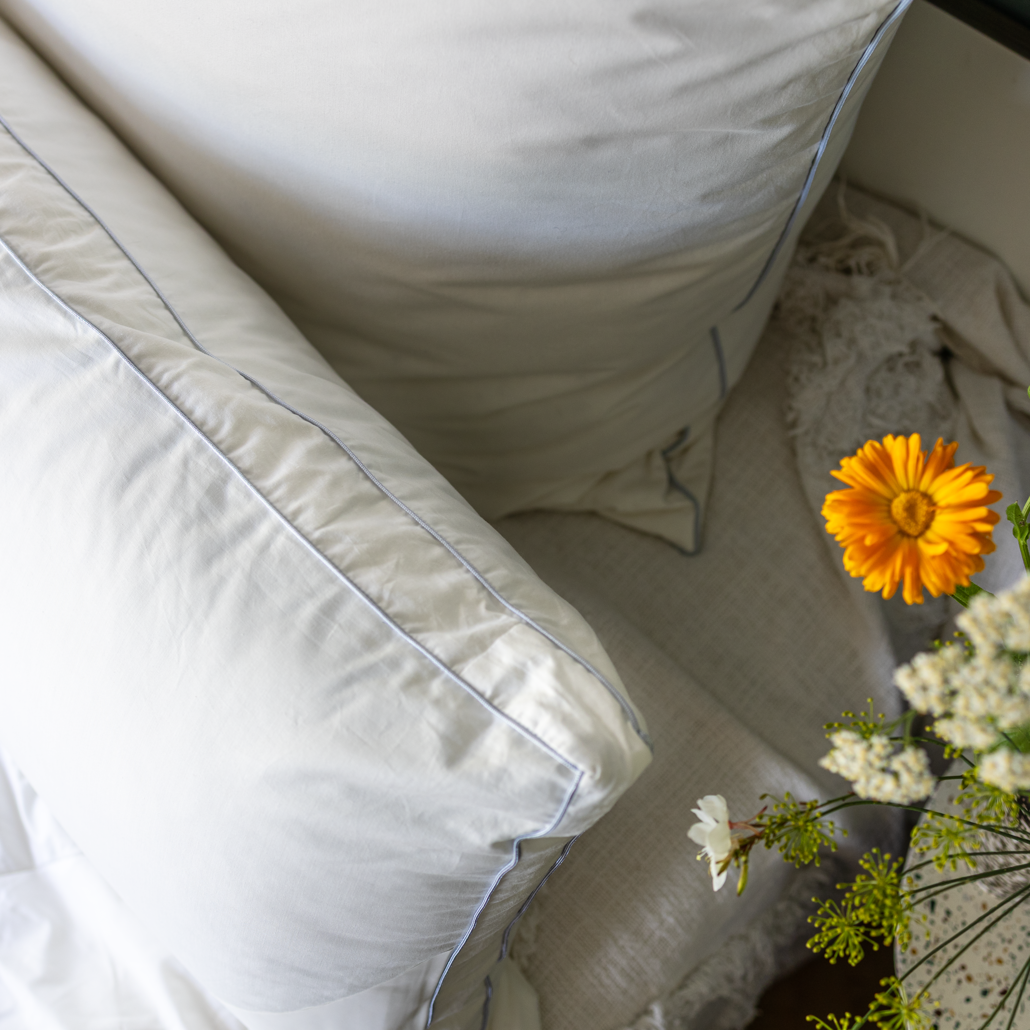 Zwei Paradies Kissen mit feiner blauer Flechtbiese auf einem hellen Stoff, daneben ein Strauß mit orangefarbener Blume und weißen Blüten