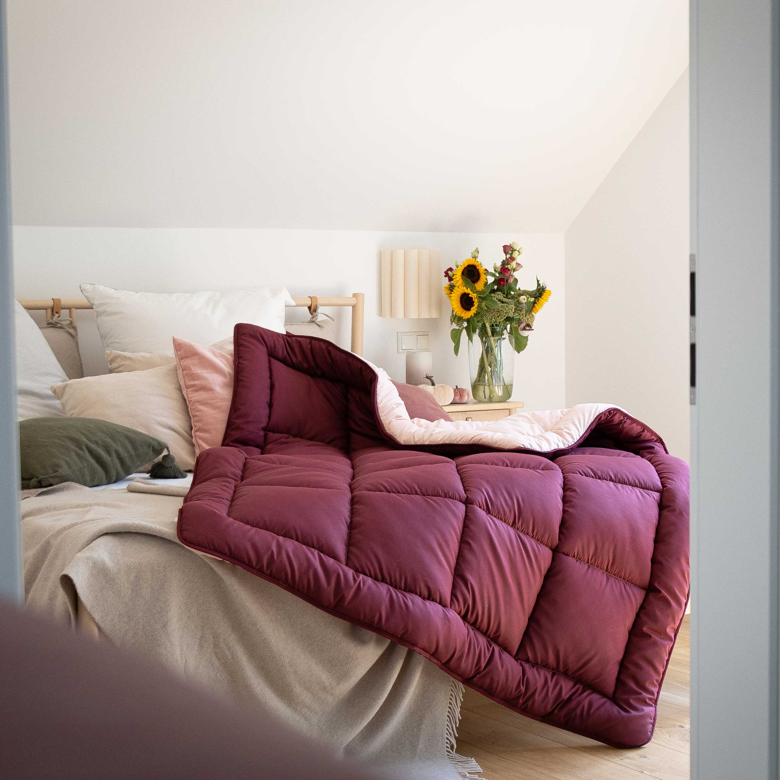 Bett mit weinroter Steppdecke, mehreren Kissen und einem Nachttisch mit Sonnenblumen in Vase #farbe_bordeaux/abendsand