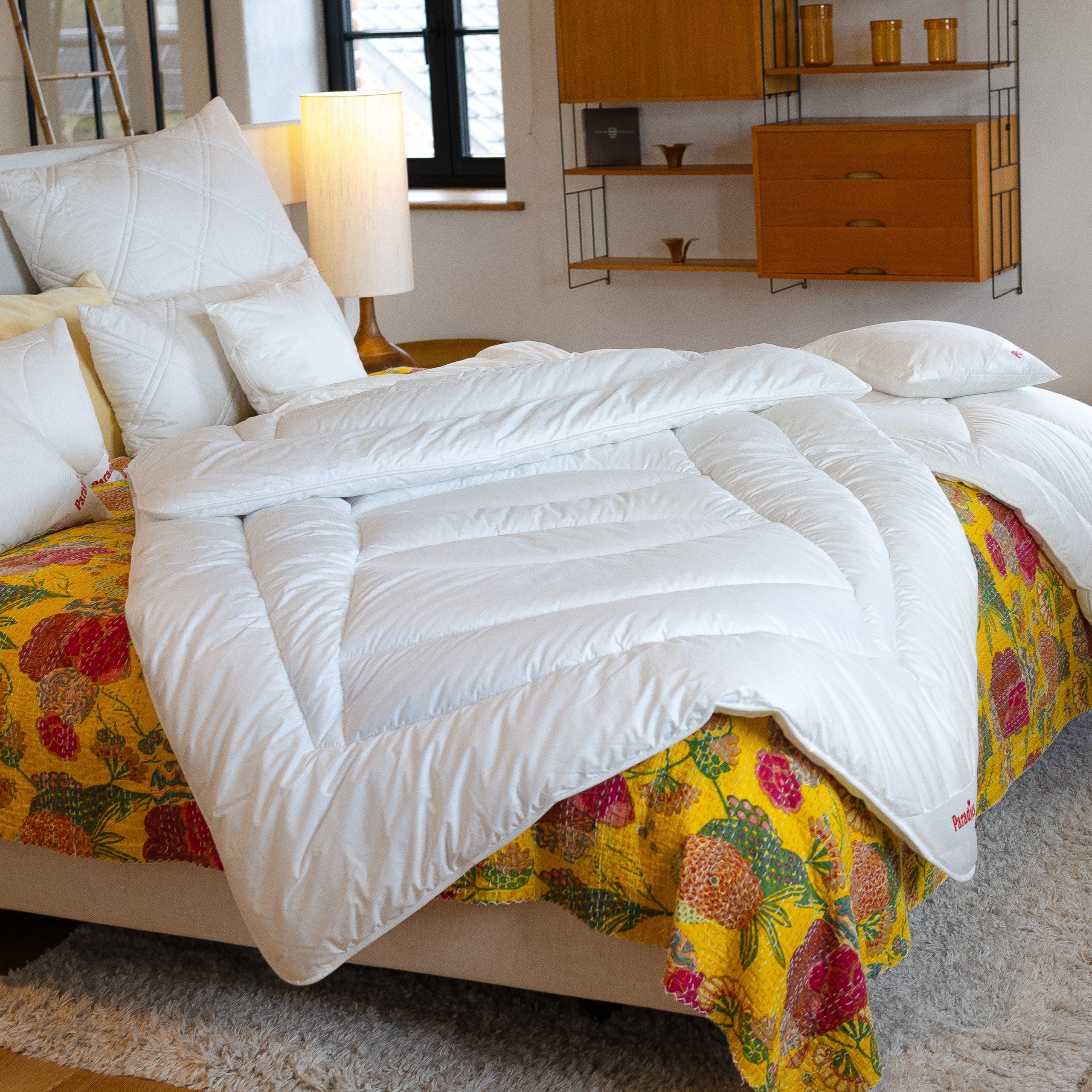 Weiße Avera Bio Ganzjahresdecke liegt auf einem Bett mit gelber, gemusterter Bettdecke in einem hellen Schlafzimmer mit Holzregal und Lampe.