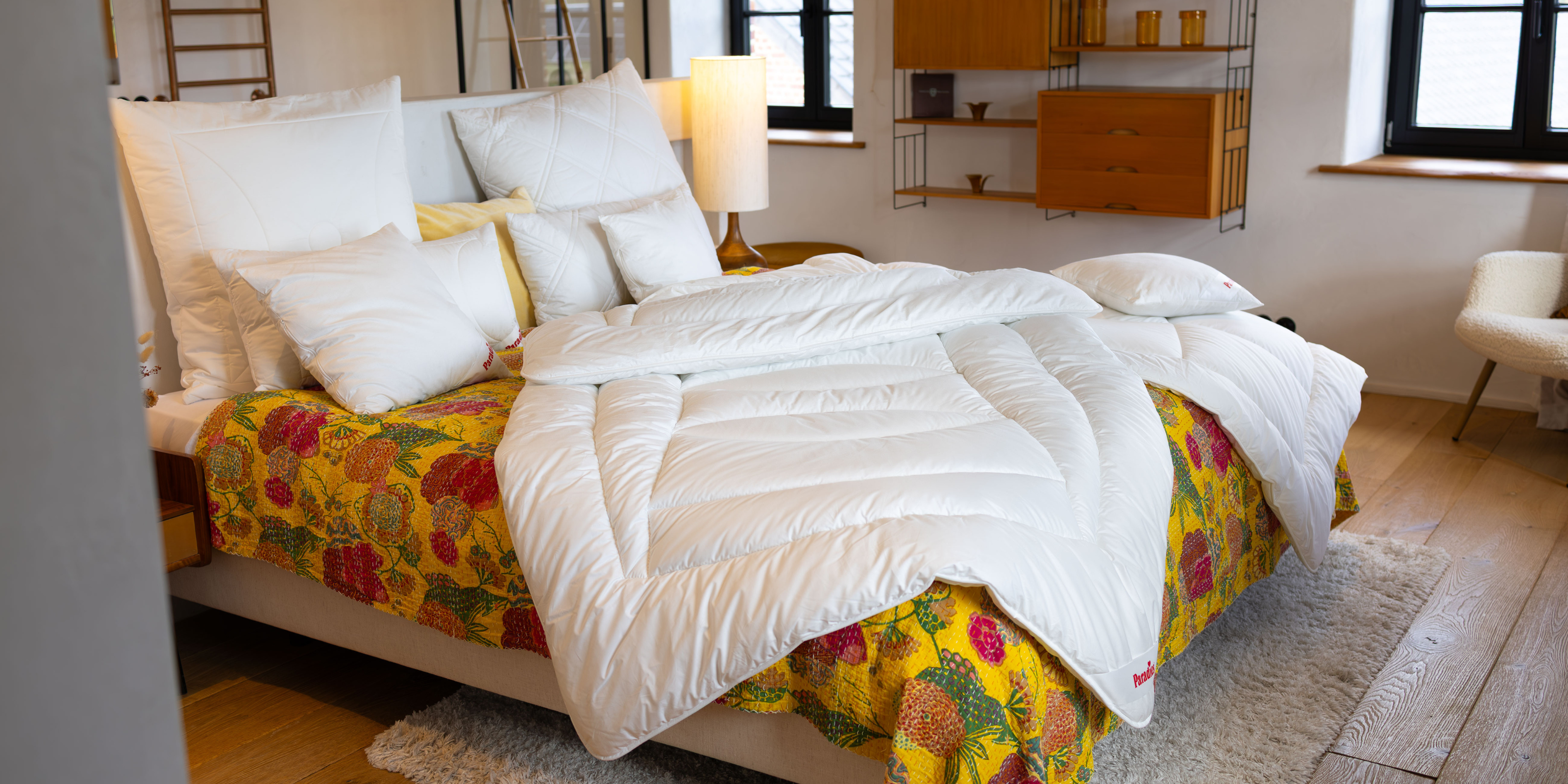 Einer heller, morderner Raum mit schön strukturiertem Holzfußboden. Auf dem Bett liegt eine gelb-geblümte Tagesdecke, die an frische Fühlings- oder Sommertage erinnert. Oben drauf unsere Ganzjahresbettdecke Avera mit oval-geschwungenen Steppnähten und einem weißen Bezug aus reiner Biobaumwolle. 