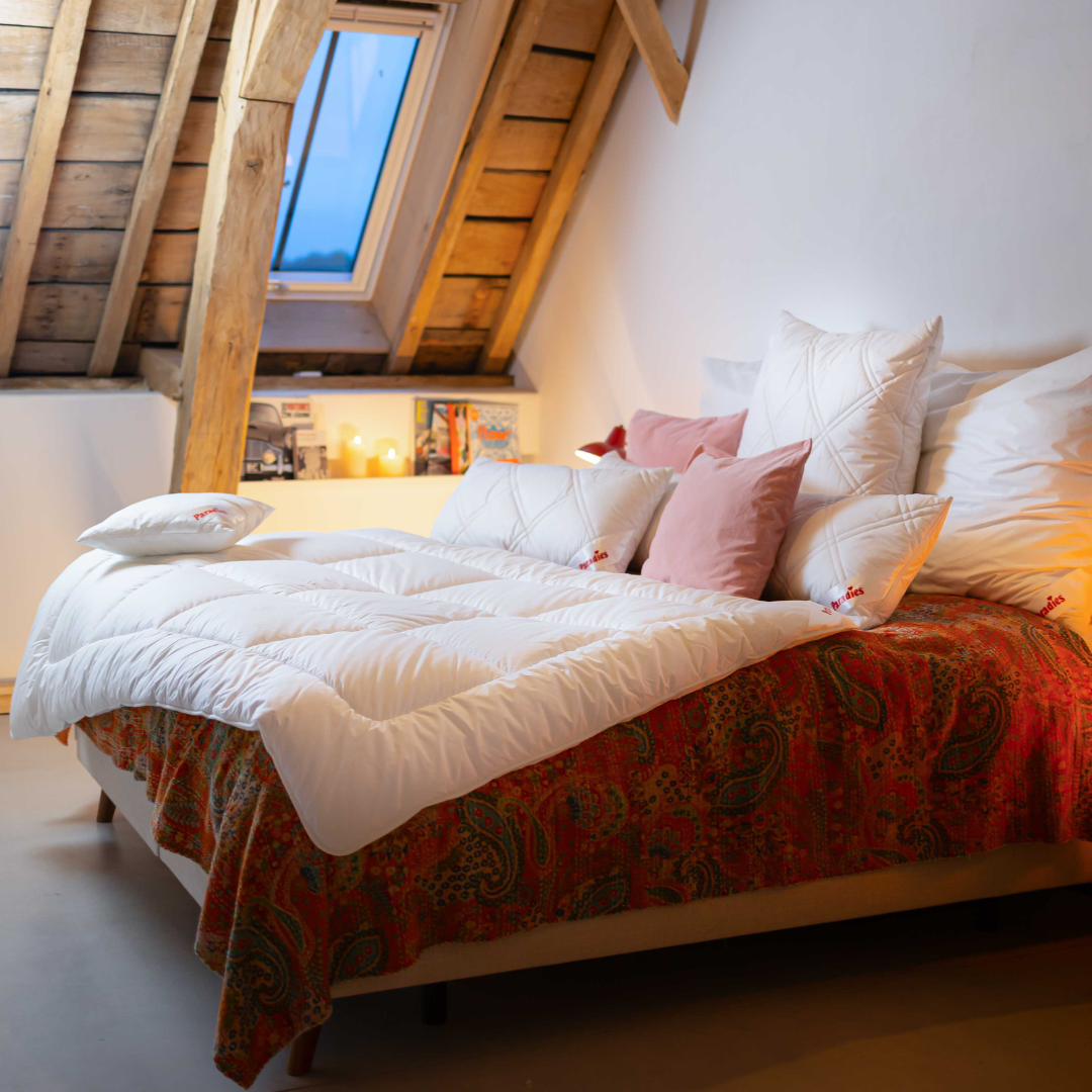 Bett mit weißer Winterbettdecke und mehreren Kissen unter einem Dachfenster in einem Dachzimmer