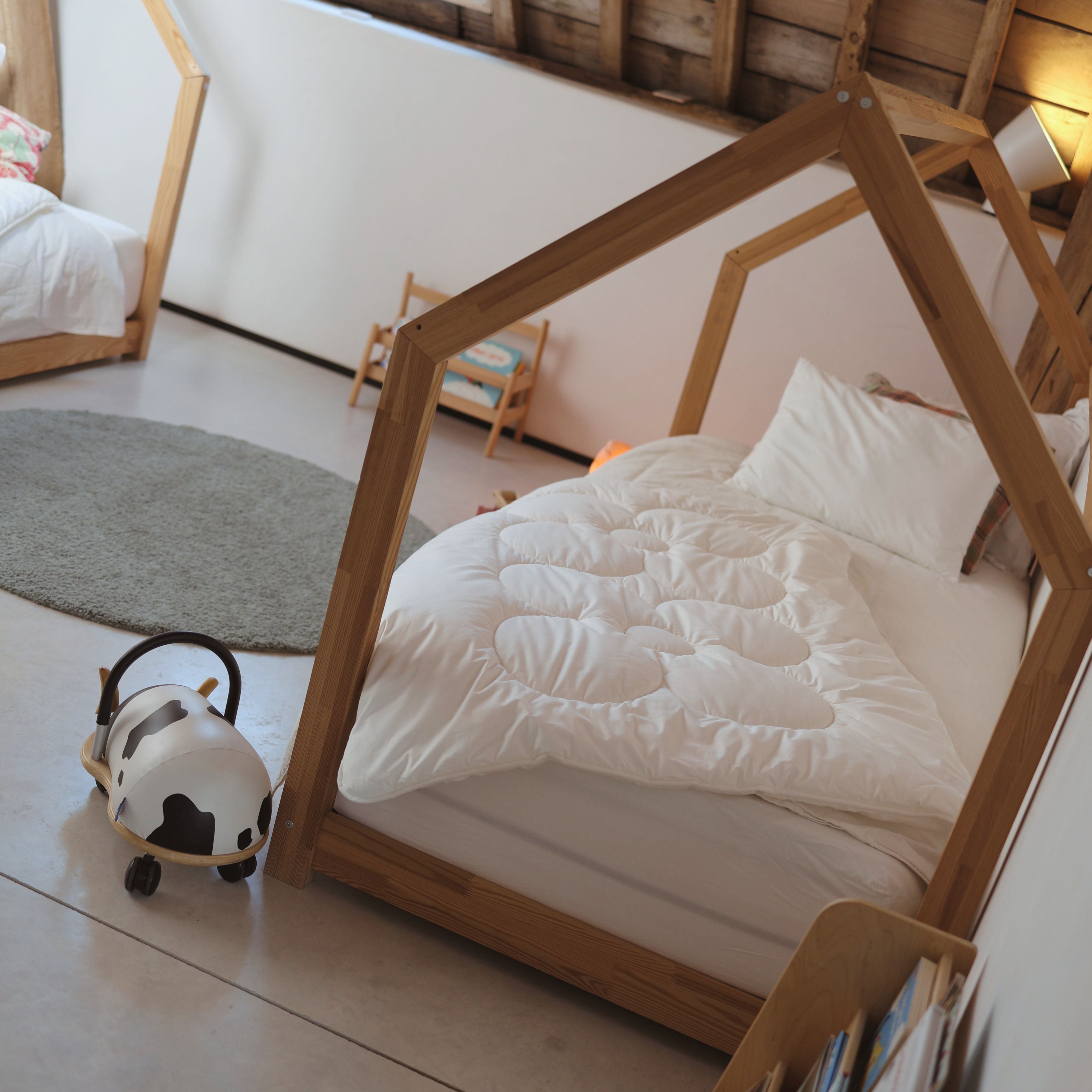 Kinderbett mit Holzrahmen in Hausform, weißer Bettdecke und Kissen neben rundem Teppich und Kinderspielzeug