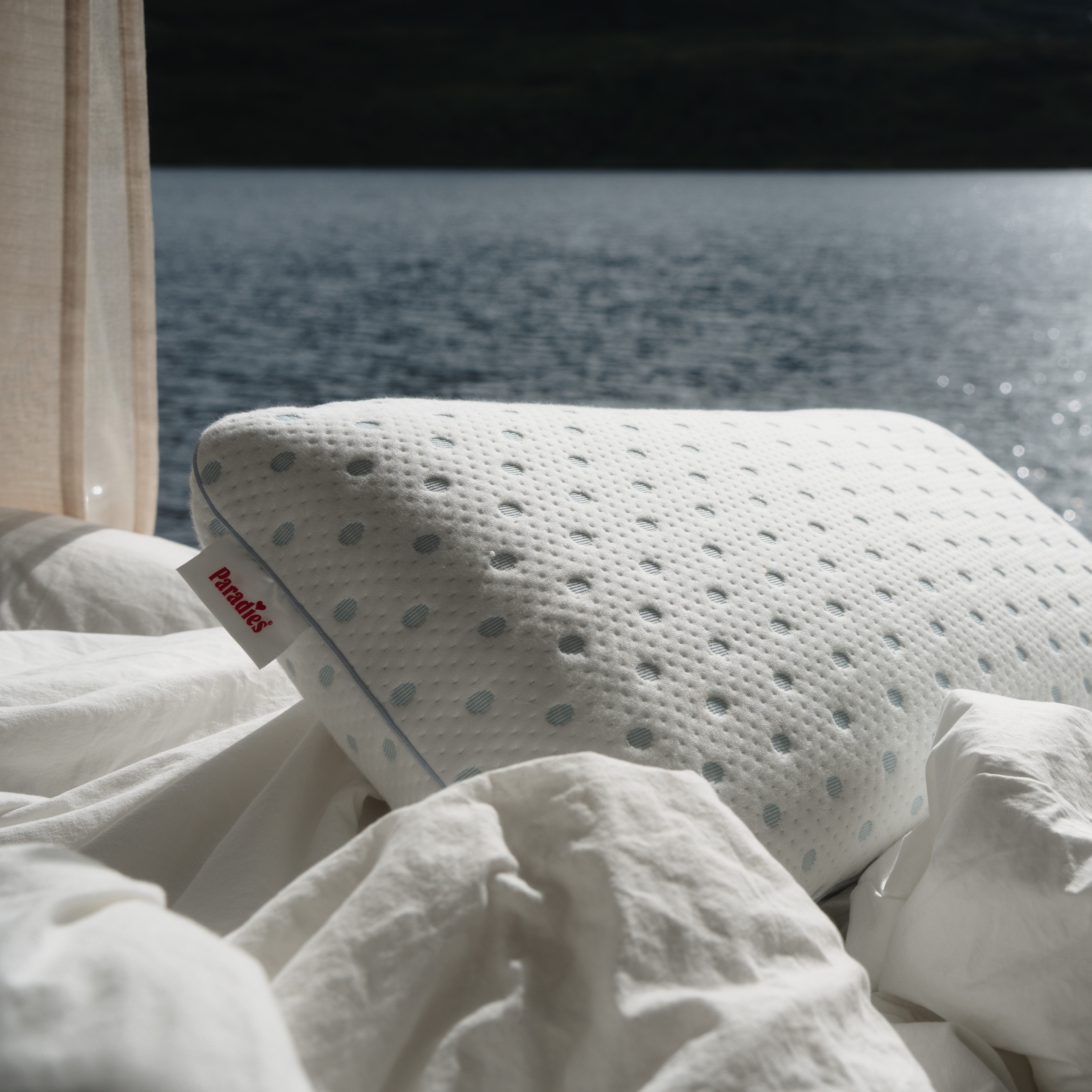 Das weiße Kissen Flexogel Dream mit blauen Punkten liegt auf einem Bett mit weißen Laken. Am Kissenrand steht Paradies. Im Hintergrund ist ein See zu sehen.