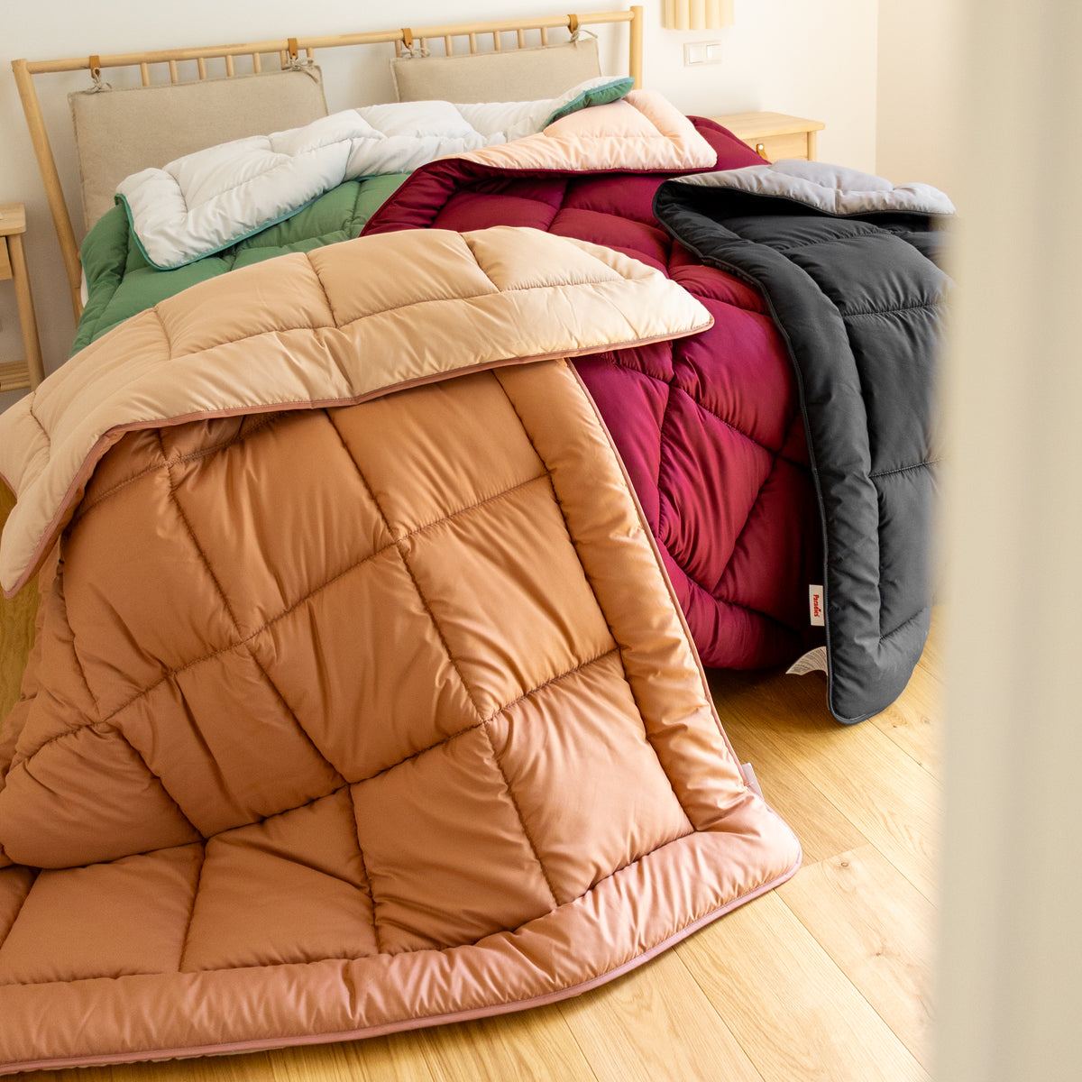 Vier auf einem Bett liegende, gefaltete Steppdecken in Beige, Dunkelrot, Grün und Schwarz
