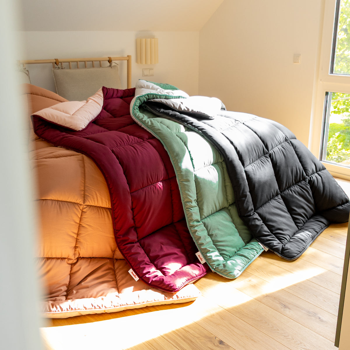 Vier unterschiedlich farbige Steppdecken hängen von einem Bett mit Holzlattenkopfteil auf einen hellen Holzboden herab
