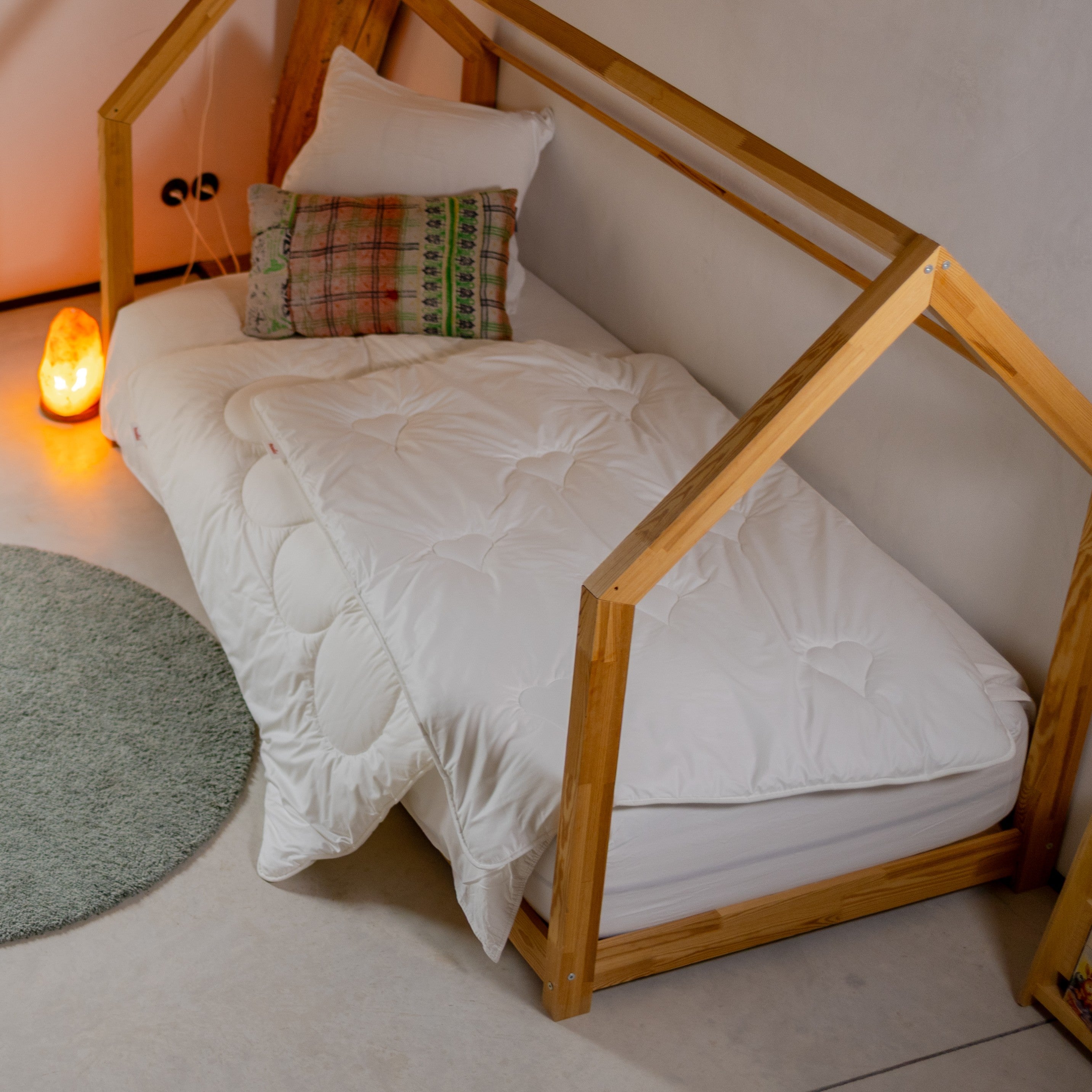 Kinderbett mit Holzrahmen in Hausform, weißer Bettdecke und zwei Kissen neben rundem Teppich und Salzlampe