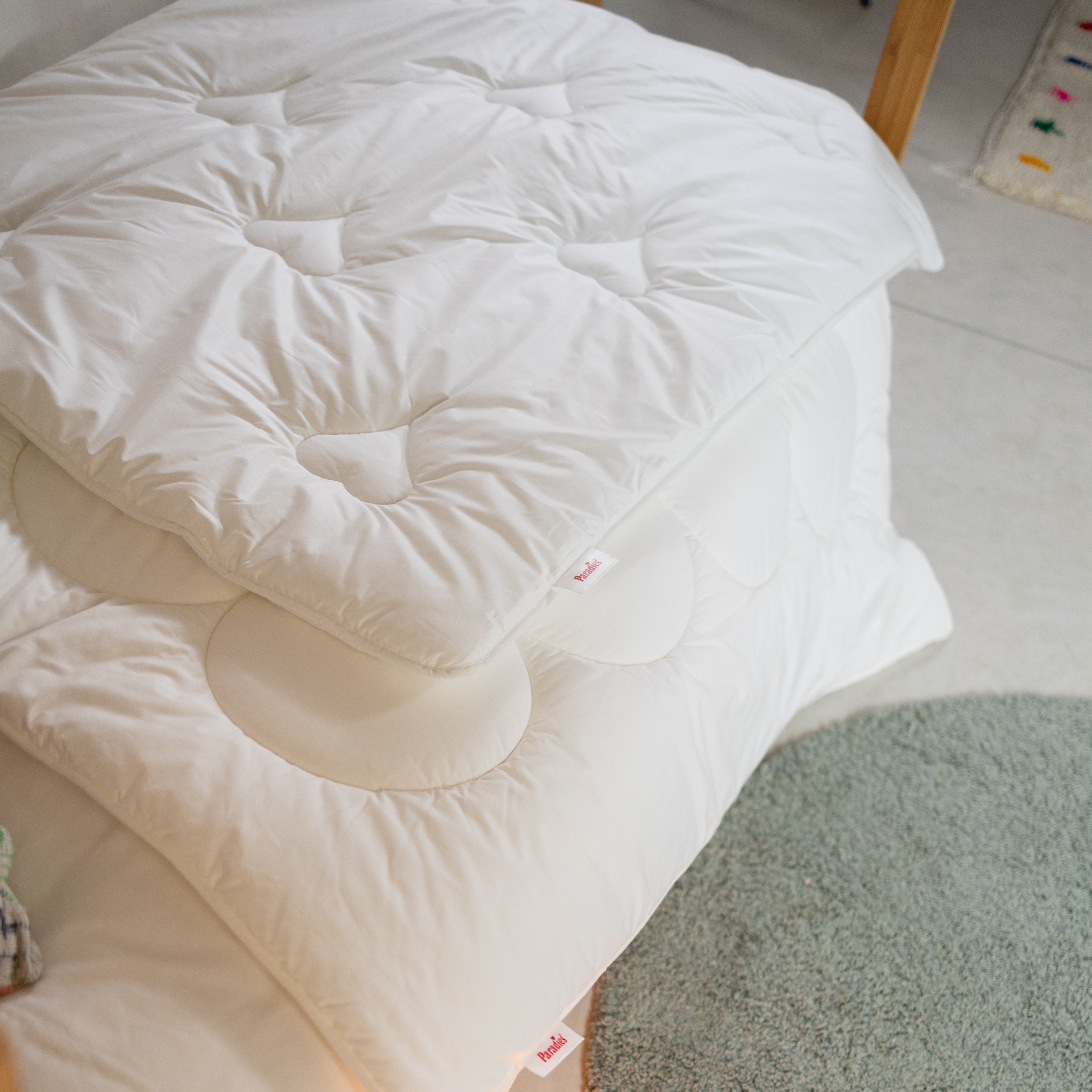 Zwei übereinandergelegte weiße Paradies Steppdecken auf einem Bett neben einem runden grauen Teppich