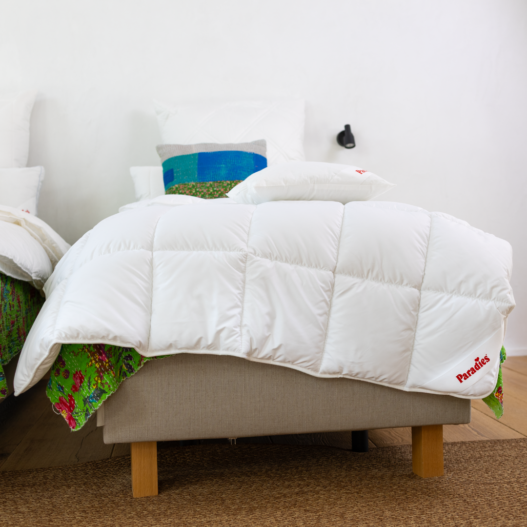 Bett mit weißer Steppdecke, die an der Ecke den roten Schriftzug 'Paradies' trägt, und buntem Kissen im Hintergrund