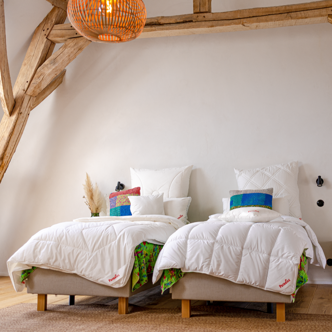 Zwei Einzelbetten stehen nebeneinander in einer alten, umgebauten Scheune. An der Decke sieht man noch die alten Holzbalken. Die den Betten liegt jeweils eine Mischung aus bunt-geblümten Tagesdecken und farbenfrohen Kissen, gepaart mit einer Mischung verschiedener weißer Paradies Kopfkissen und Textilfasedrbettdecken mit variablen Steppmustern.