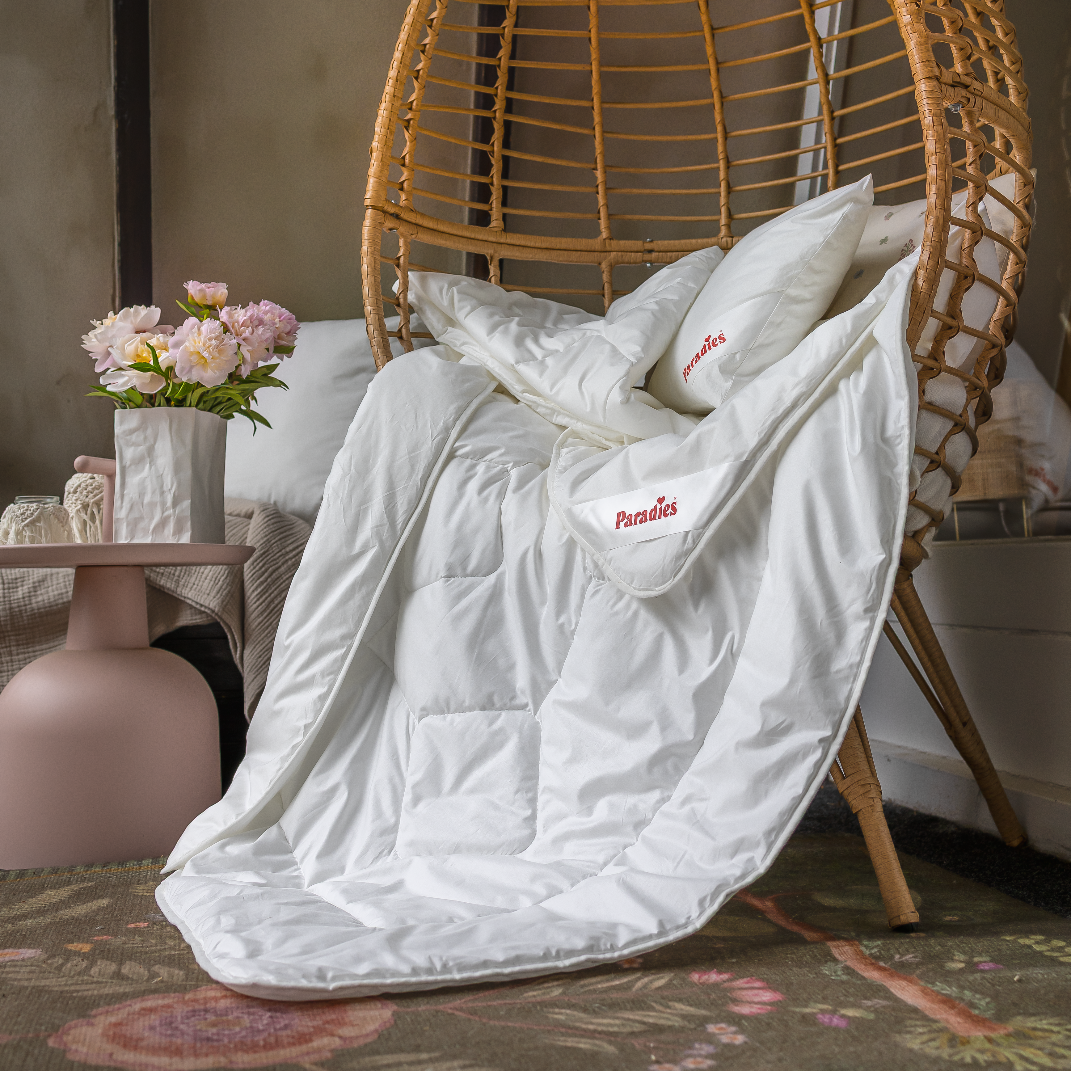 Weiße Prima Bio Sommerbettdecke, drapiert auf einem geflochtenen Stuhl neben einem Tisch mit Vase und Blumen