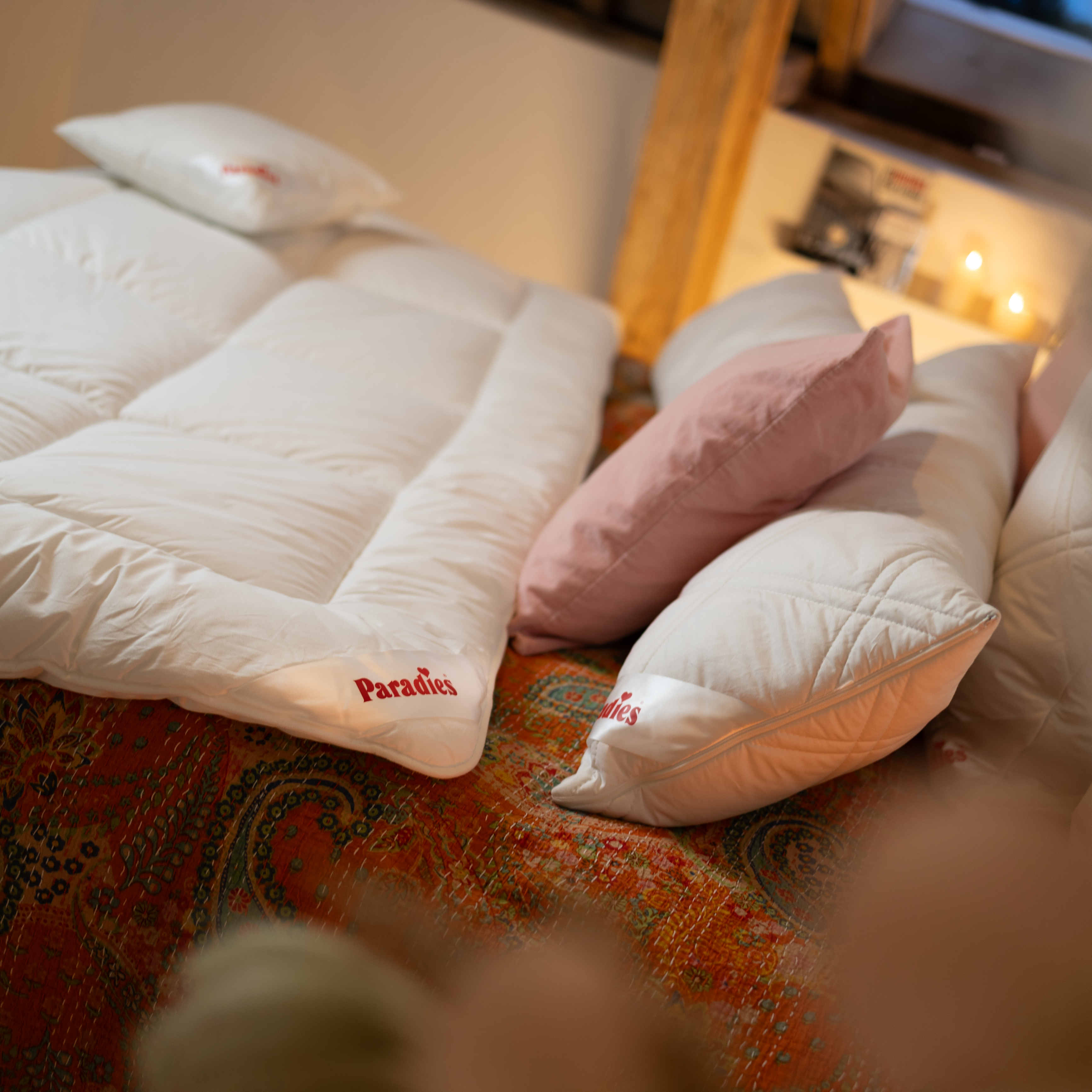 Bett mit weißer Steppdecke, die an der Ecke den roten Schriftzug 'Paradies' trägt, und buntem Kissen im Hintergrund